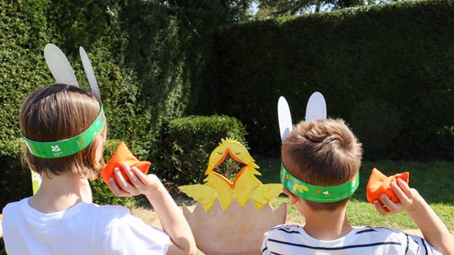 Two people with their backs to the camera holding an orange bean bag ready to throw at a wooden chick with it's mouth open.